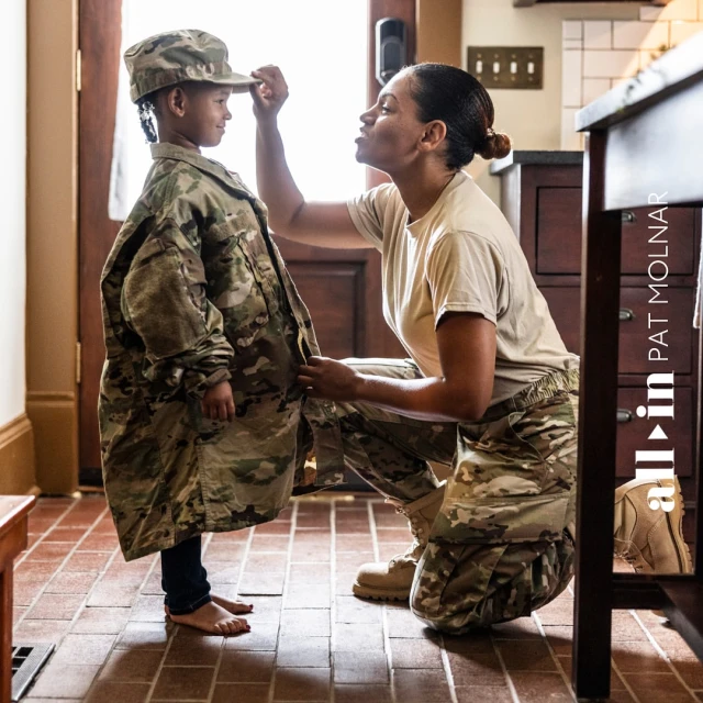 A mother, kneeling, adjusts the hat of a young child in an oversized military uniform inside a home with warm lighting.
