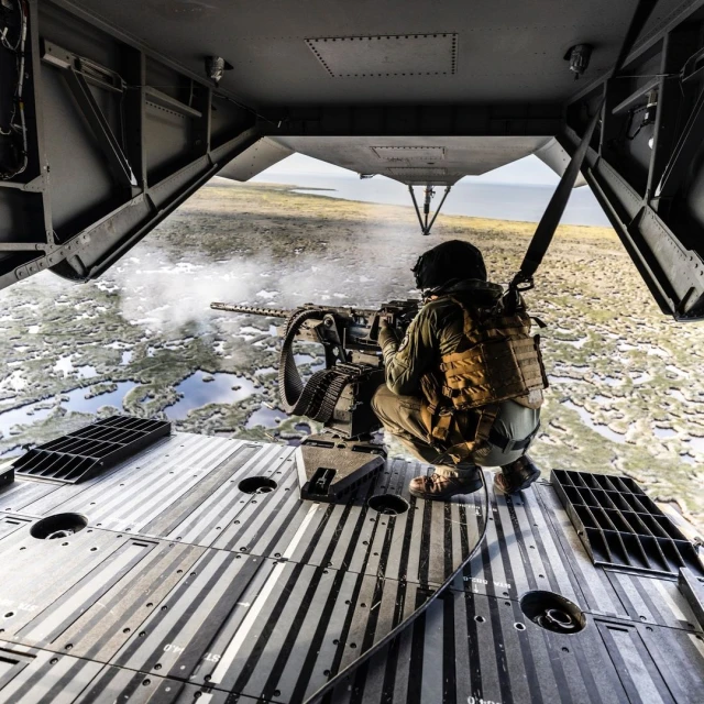 Military personnel firing a machine gun from the open back of a helicopter, with a landscape of water and land visible below.