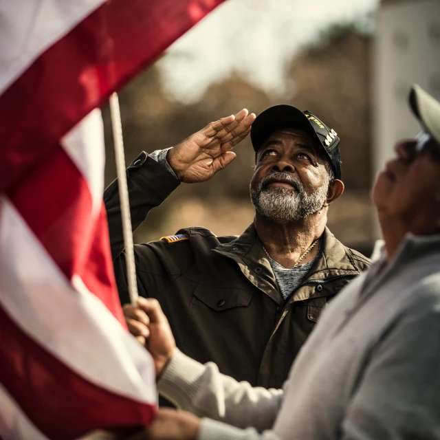 Two men salute a waving American flag outdoors in a patriotic display of respect and remembrance on a sunny day.