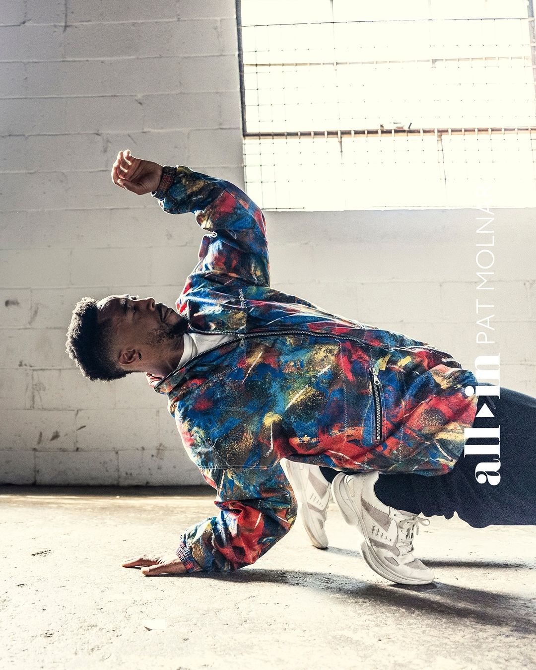 Male dancer strikes dynamic pose in industrial space. Wearing colorful jacket, dark pants, white shoes, he's captured in mid-movement.