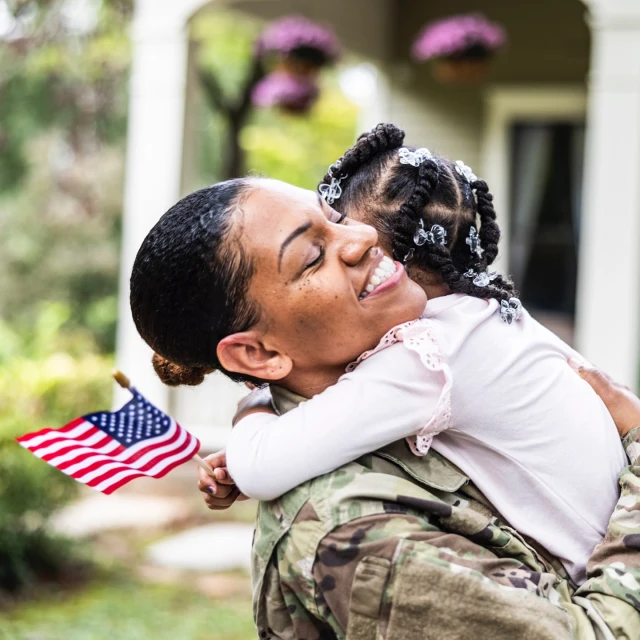 A military mother in uniform hugging her young daughter outside their home, while holding a small American flag, smiling, family moment.
