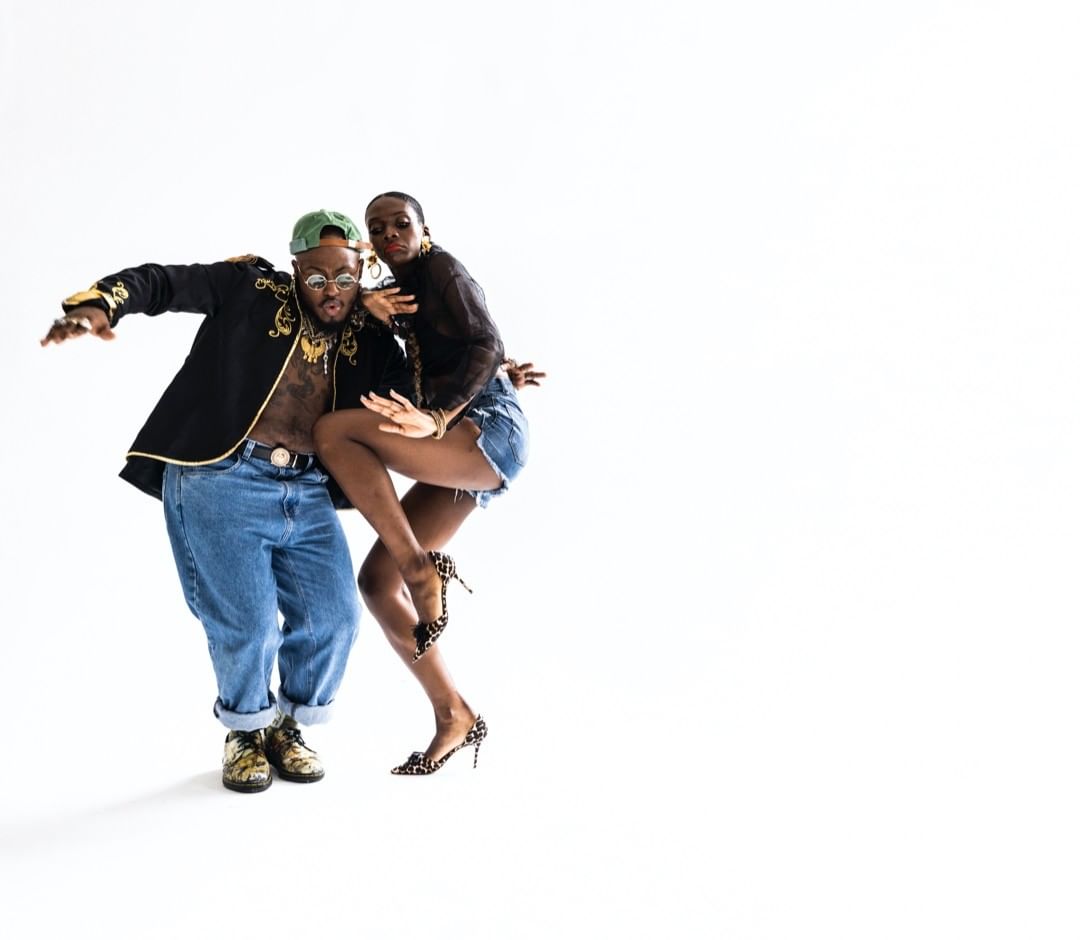 Two people, an adult man and woman, pose in a dance position. The man is wearing a jacket, jeans, and cap, while the woman wears a black blouse and heels.
