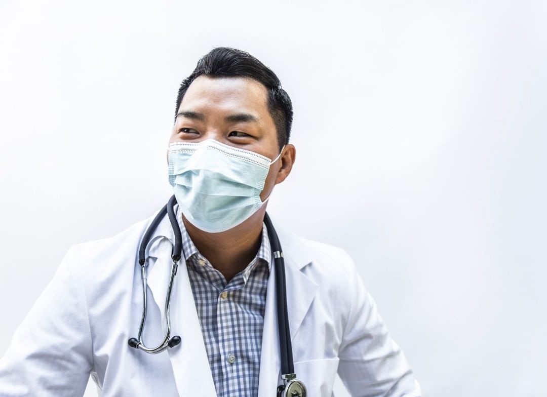 A young Asian male doctor in a white coat and surgical mask smiles in a bright indoor setting, wearing a stethoscope and plaid shirt.