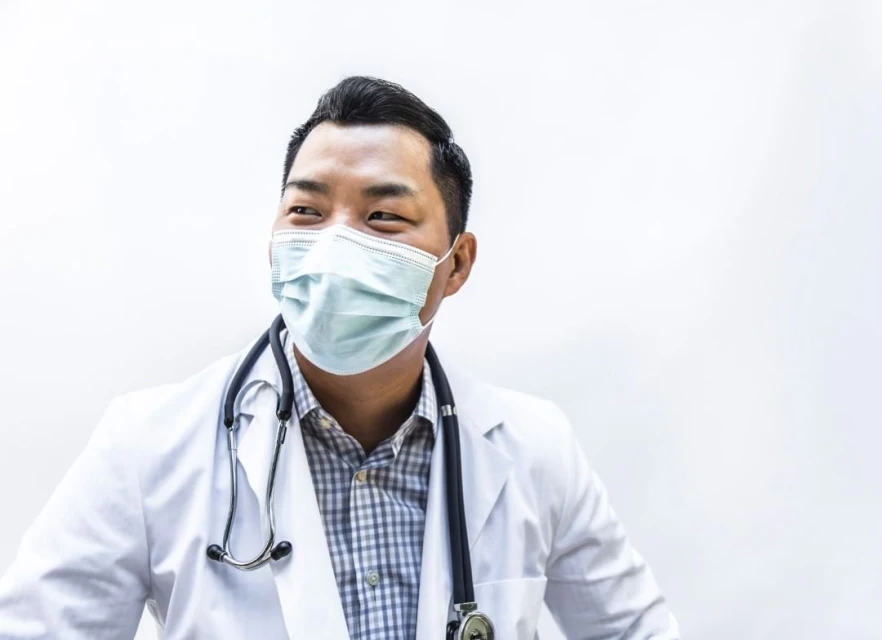 A young Asian male doctor in a white coat and surgical mask smiles in a bright indoor setting, wearing a stethoscope and plaid shirt.