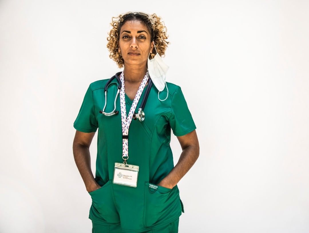 Medical professional in green scrubs with stethoscope and face mask around neck, poses against a plain, light background in a portrait.