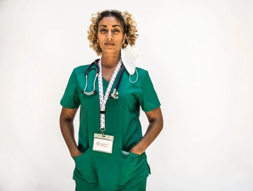 Medical professional in green scrubs with stethoscope and face mask around neck, poses against a plain, light background in a portrait.