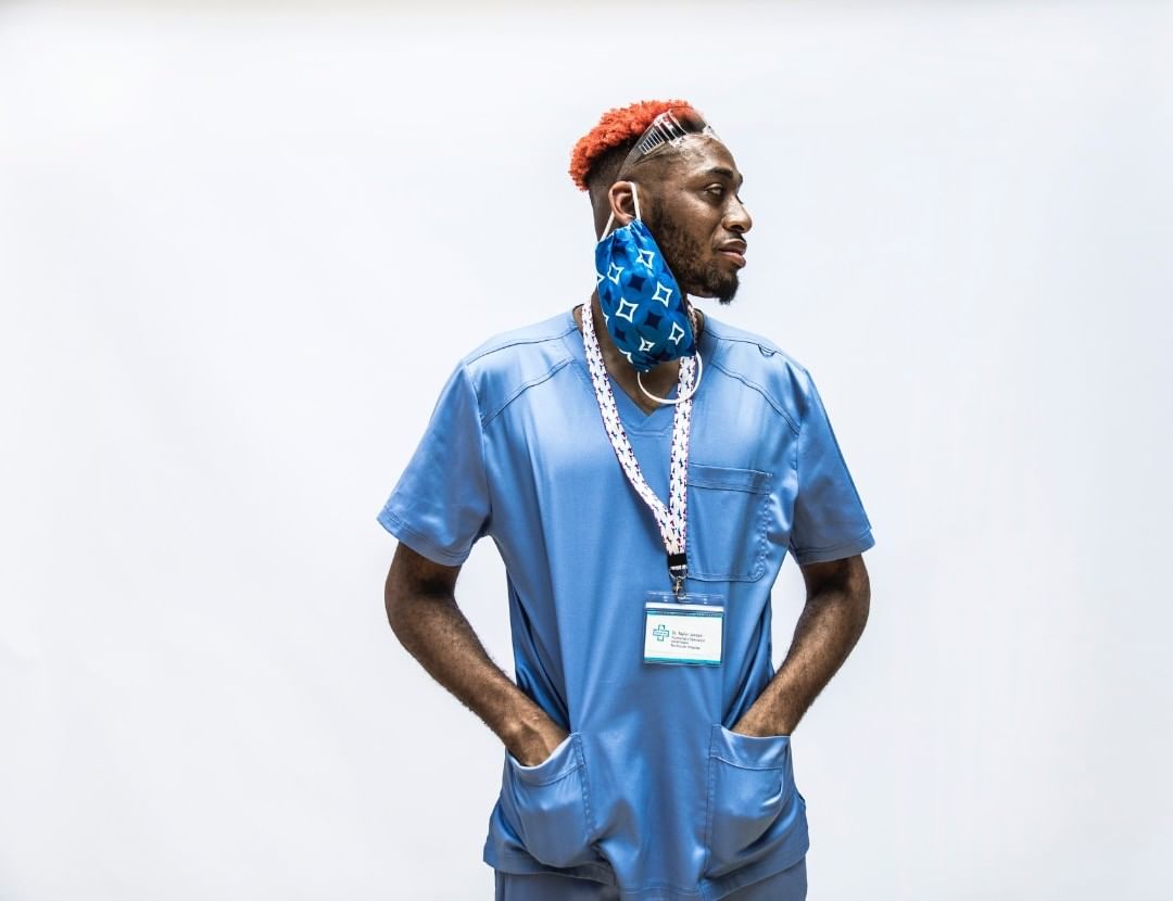 An African-American healthcare worker with orange hair, wearing a blue scrub suit with a face mask around the neck, stands in a studio.