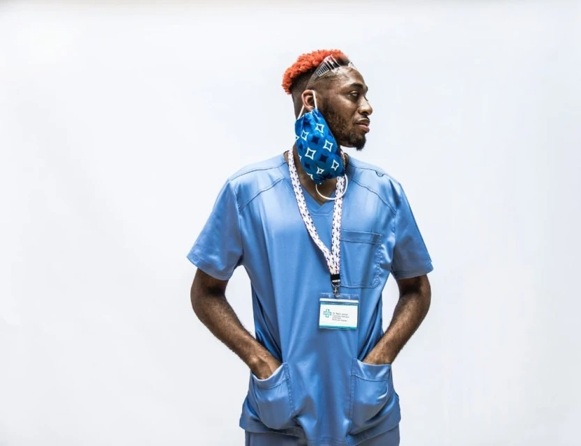 An African-American healthcare worker with orange hair, wearing a blue scrub suit with a face mask around the neck, stands in a studio.
