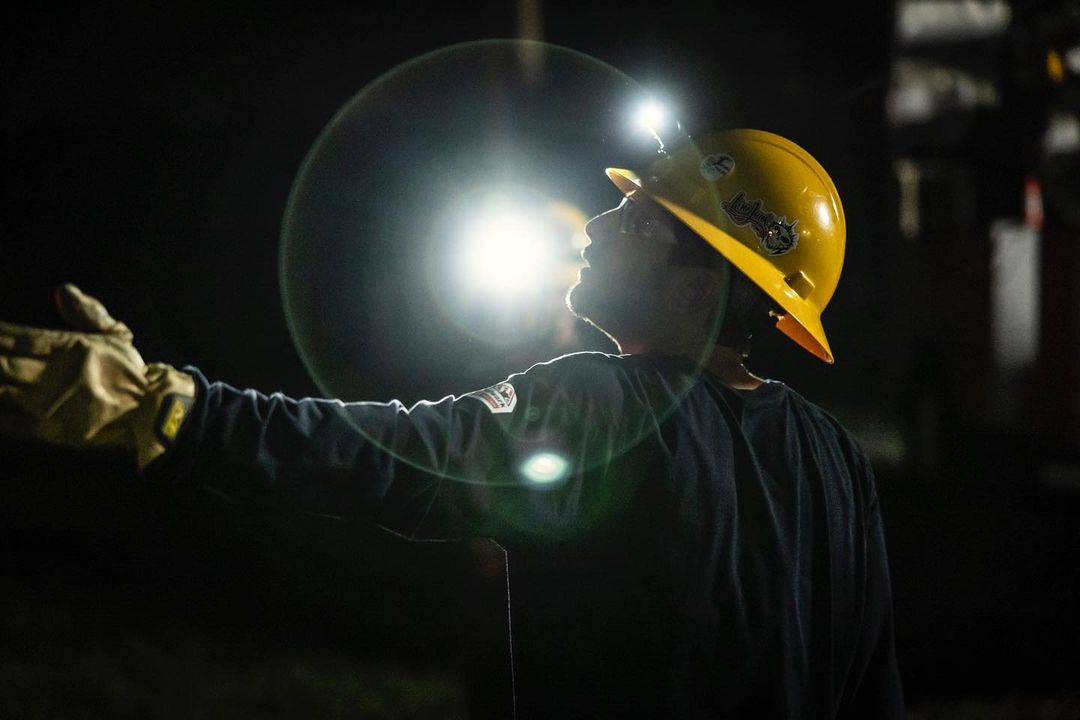 Construction worker in hardhat and gloves, illuminated by headlamp, raises hand in dark setting with some equipment in background.