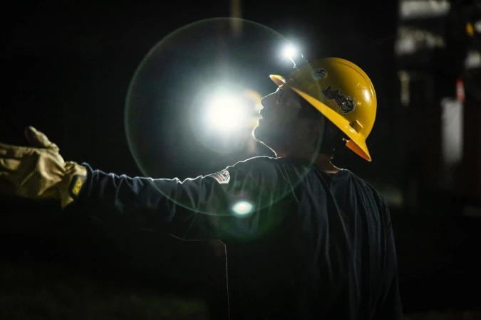 Construction worker in hardhat and gloves, illuminated by headlamp, raises hand in dark setting with some equipment in background.