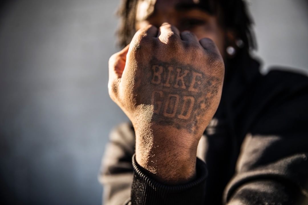 Close up of an adult with dreadlocks and a tattoo on their fist that reads 'Bike God', posing outdoors with grey background.