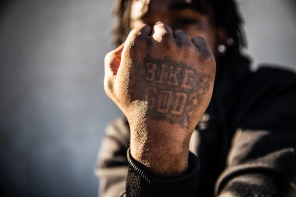 Close up of an adult with dreadlocks and a tattoo on their fist that reads 'Bike God', posing outdoors with grey background.