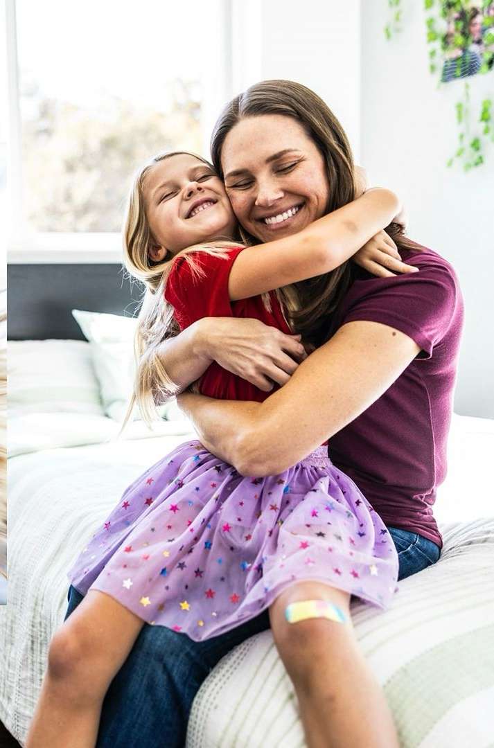 A mother and daughter embrace indoors; the young girl has a purple skirt and a bandage on her leg. They are smiling and happy.