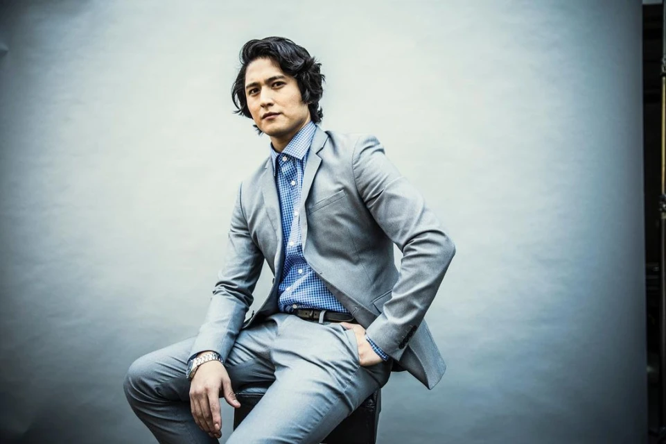 Adult male in grey suit and blue shirt sits on a black stool in a photo studio, posing for portrait with hand in pocket.