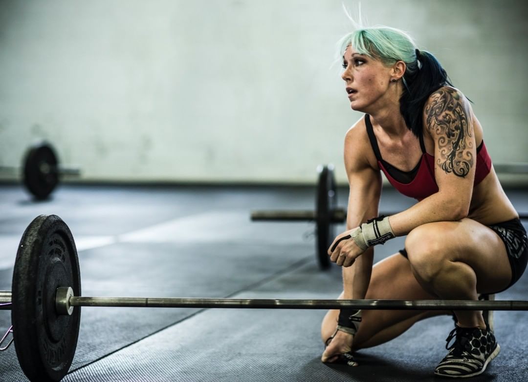Fit woman with tattoos and colorful hair resting between sets with barbell on the floor in an indoor gym. Sport and fitness scene.