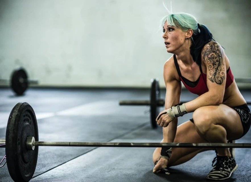 Fit woman with tattoos and colorful hair resting between sets with barbell on the floor in an indoor gym. Sport and fitness scene.