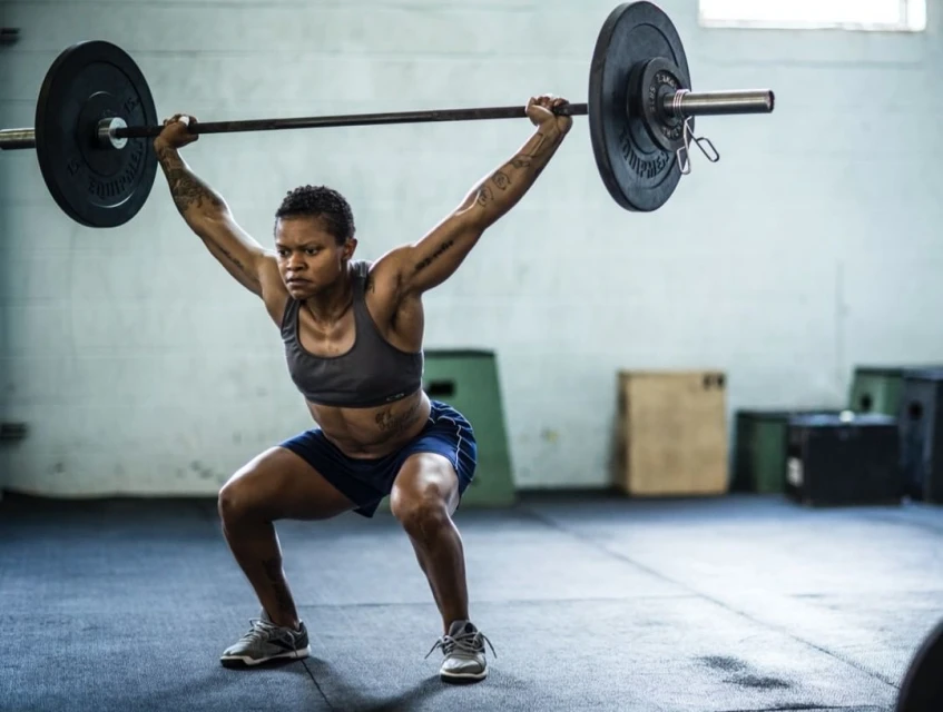 A strong woman powerfully performs a barbell overhead squat in a bright fitness studio, showcasing strength and determination in her workout.