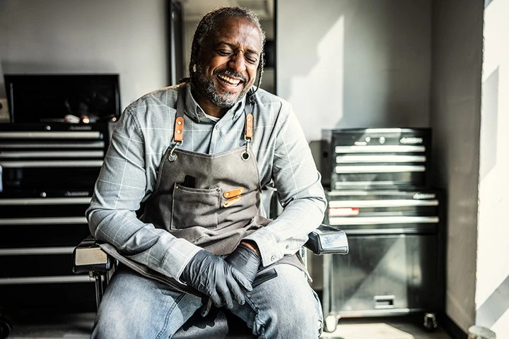 An African American barber with beard and grey hair laughing while wearing an apron and gloves, sitting in a barber's chair.