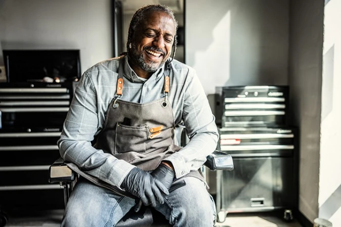 An African American barber with beard and grey hair laughing while wearing an apron and gloves, sitting in a barber's chair.