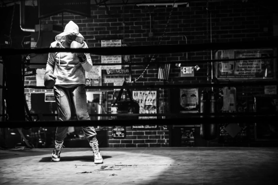 Monochrome shot of a hooded boxer with gloves in a boxing ring under a spotlight with a brick wall and signage in the background.