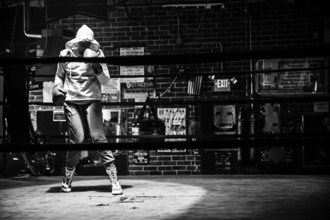 Monochrome shot of a hooded boxer with gloves in a boxing ring under a spotlight with a brick wall and signage in the background.