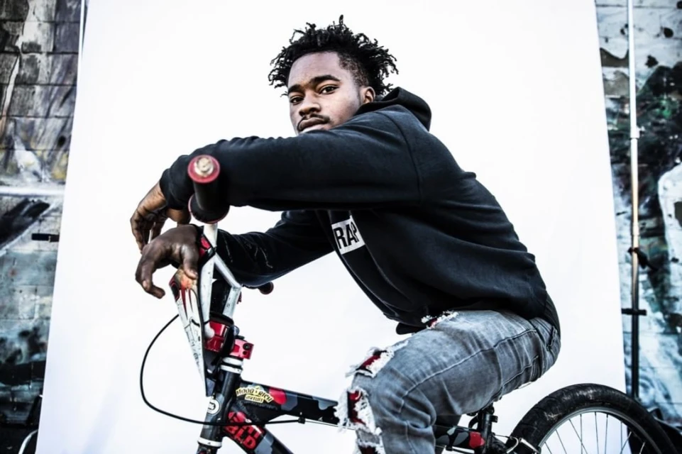 Portrait of a young Black man on a BMX bike with a dark hooded sweatshirt, sitting against a white backdrop with graffitied wall.