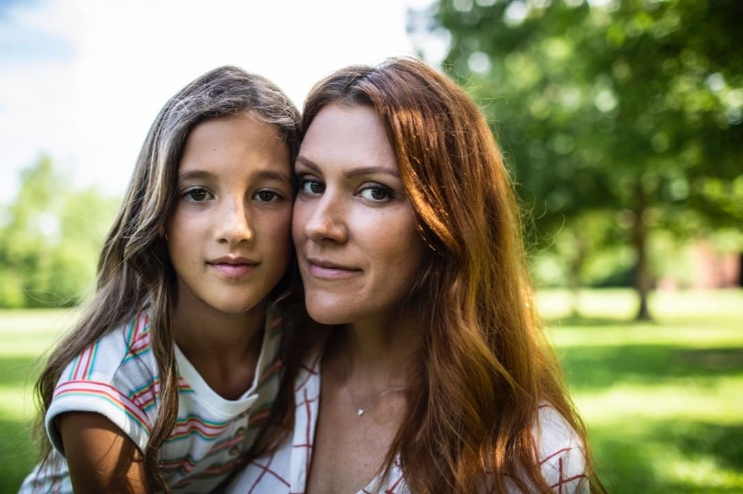 Portrait of a mother and daughter outdoors in a park-like setting; they are looking at the camera. The background includes green grass and trees.