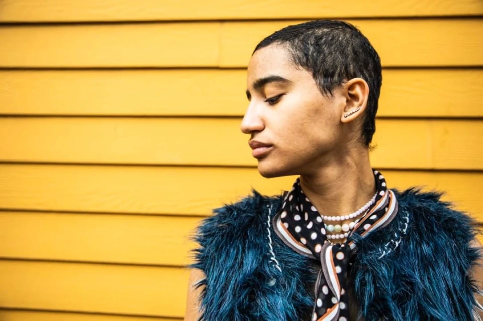 Profile of a stylish adult with shaved head wearing fur vest and pearl necklace standing against a yellow wooden wall outdoors.