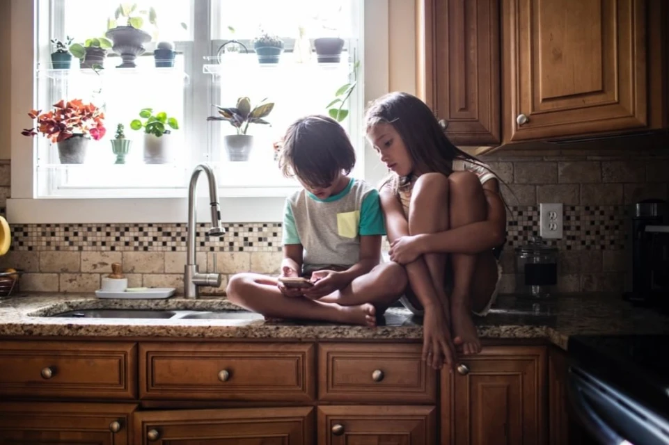 A young boy and girl sitting barefoot on a kitchen counter, looking at a cell phone, with potted plants near the window.