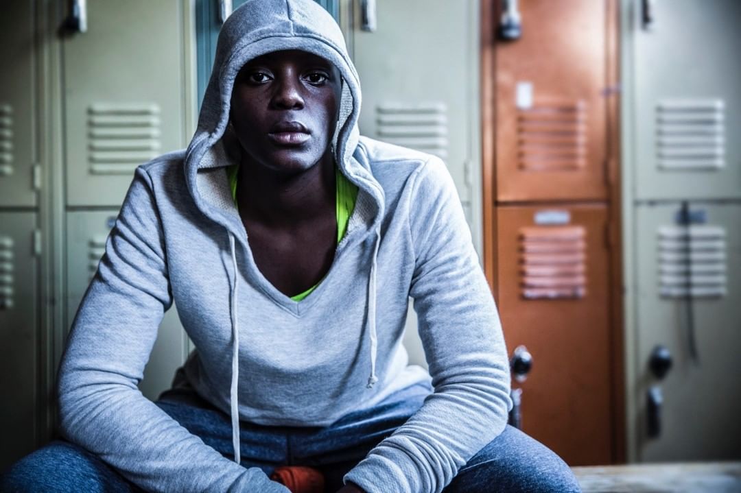 Young Black adult in a grey hoodie and blue jeans sitting in a locker room looking directly at the camera with a serious expression.