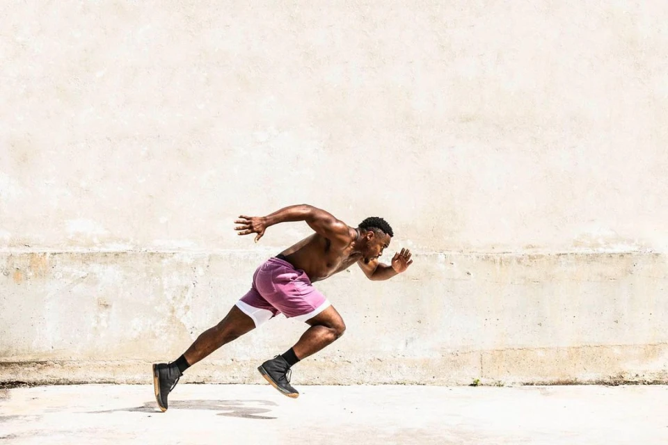 Athletic African-American man in maroon shorts, shirtless, in a sprinter's stance against a textured concrete wall, outdoor fitness.
