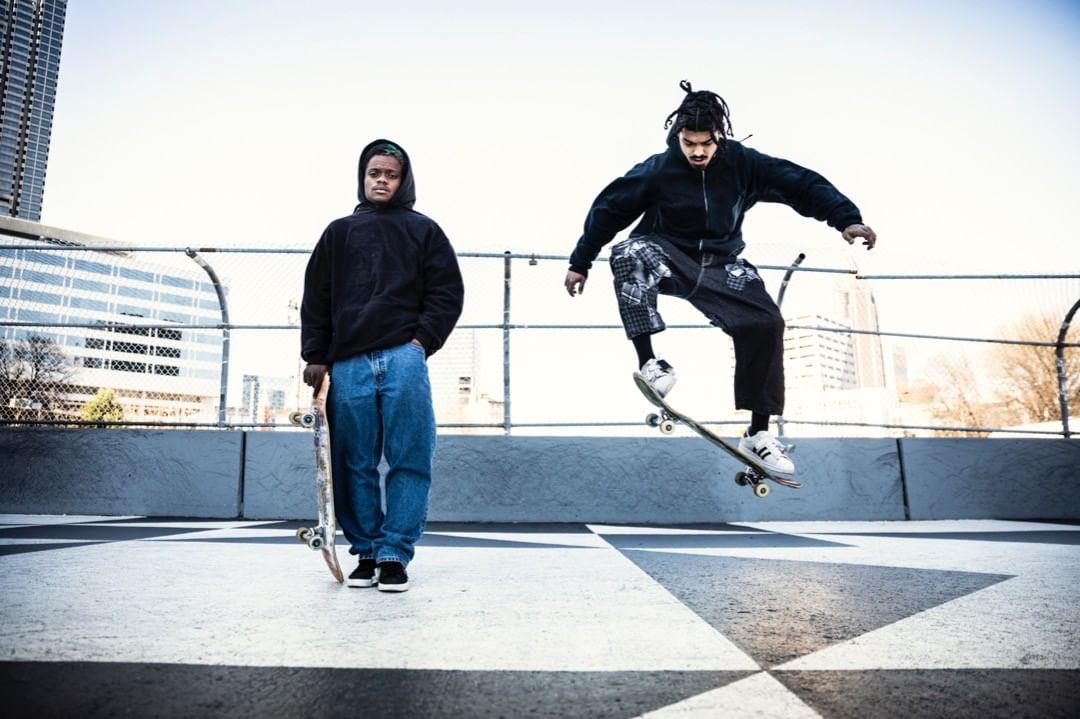 Two skateboarders, one in mid-air performing a trick, the other standing on a rooftop with a city skyline in the background.