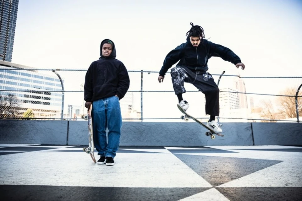 Two skateboarders, one in mid-air performing a trick, the other standing on a rooftop with a city skyline in the background.