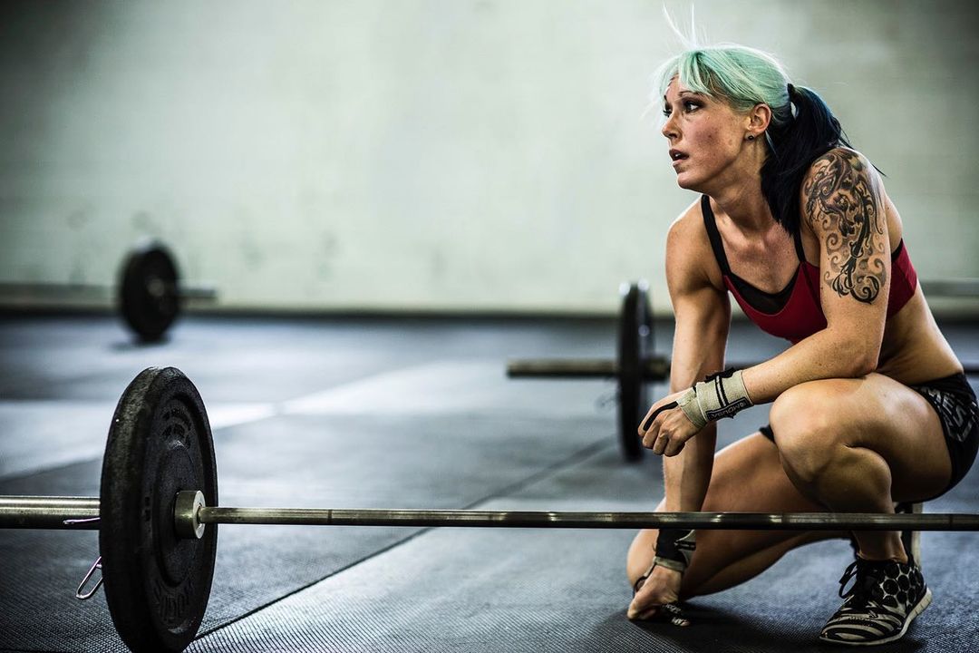 Athletic woman with blue hair rests after weightlifting in gym, wearing red sports bra and black shorts, with a barbell in foreground.