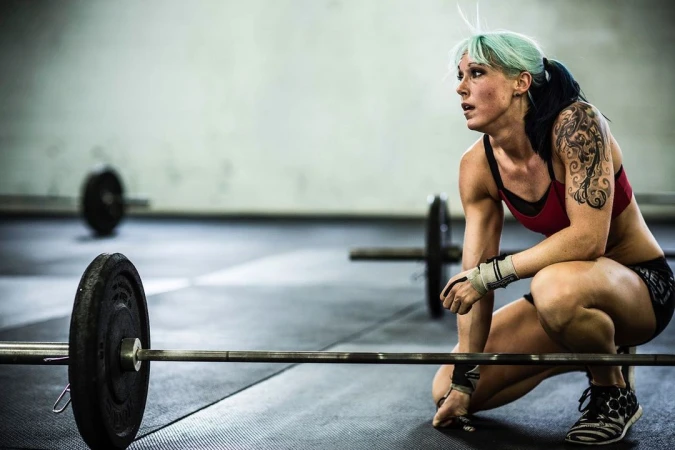 Athletic woman with blue hair rests after weightlifting in gym, wearing red sports bra and black shorts, with a barbell in foreground.