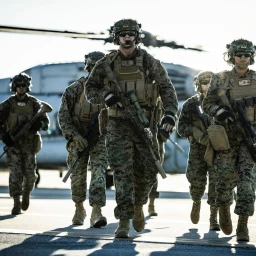 Group of armed military personnel in camouflage uniforms walking forward with a helicopter in the background