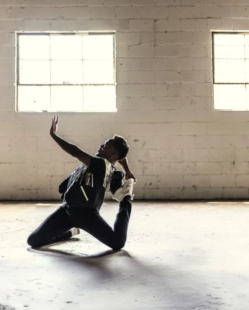 Dancer in dark clothing performing a flexible floor pose inside an industrial space with white brick walls.