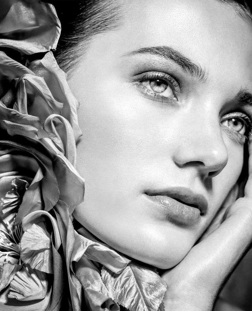 Close-up black and white beauty shot of a young woman with a silk flower accessory, soft lighting, and a contemplative expression.
