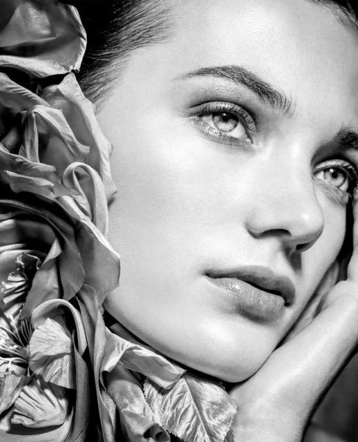 Close-up black and white beauty shot of a young woman with a silk flower accessory, soft lighting, and a contemplative expression.