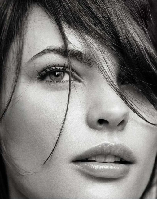 Close-up portrait of a woman with dark hair partially covering her face, emphasizing her eye, nose, and lips in black and white.