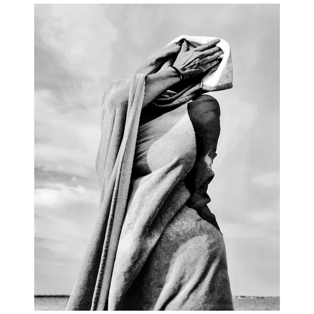 Monochrome portrait of an adult covering their face with a white towel outside with a cloudy sky and the sea in the background.