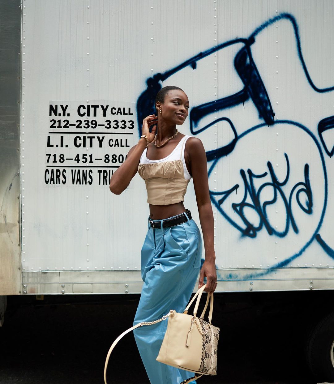 Woman in crop top, and blue pants carries a handbag in front of a commercial truck with graffiti and a phone number.