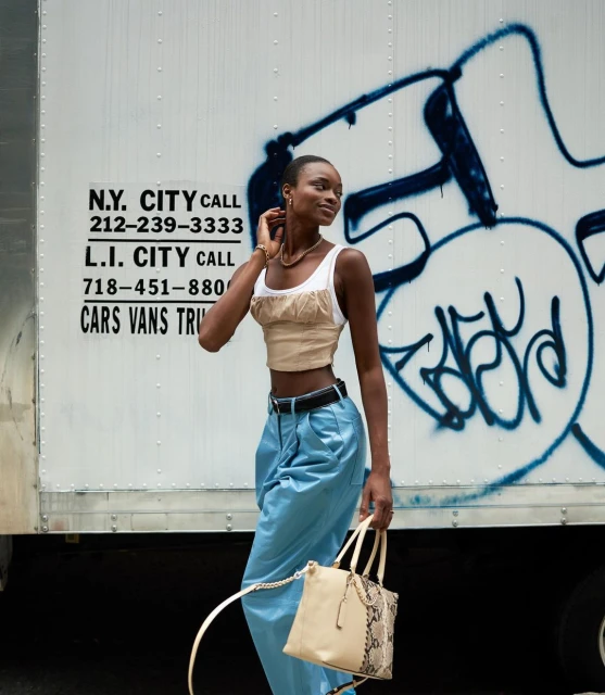 Woman in crop top, and blue pants carries a handbag in front of a commercial truck with graffiti and a phone number.