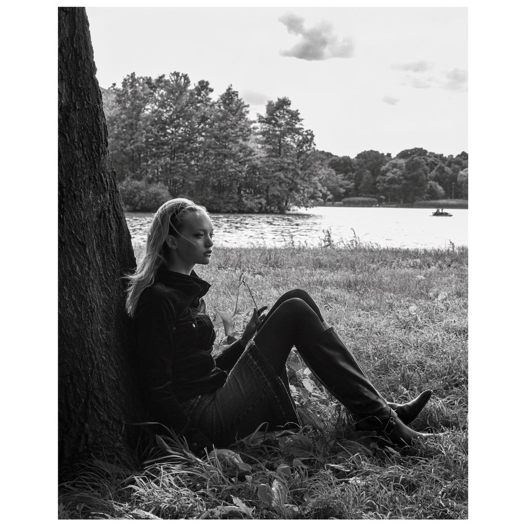 Young woman sits by tree near a lake in a black and white photograph, gazing at the water in the distance on a sunny day.