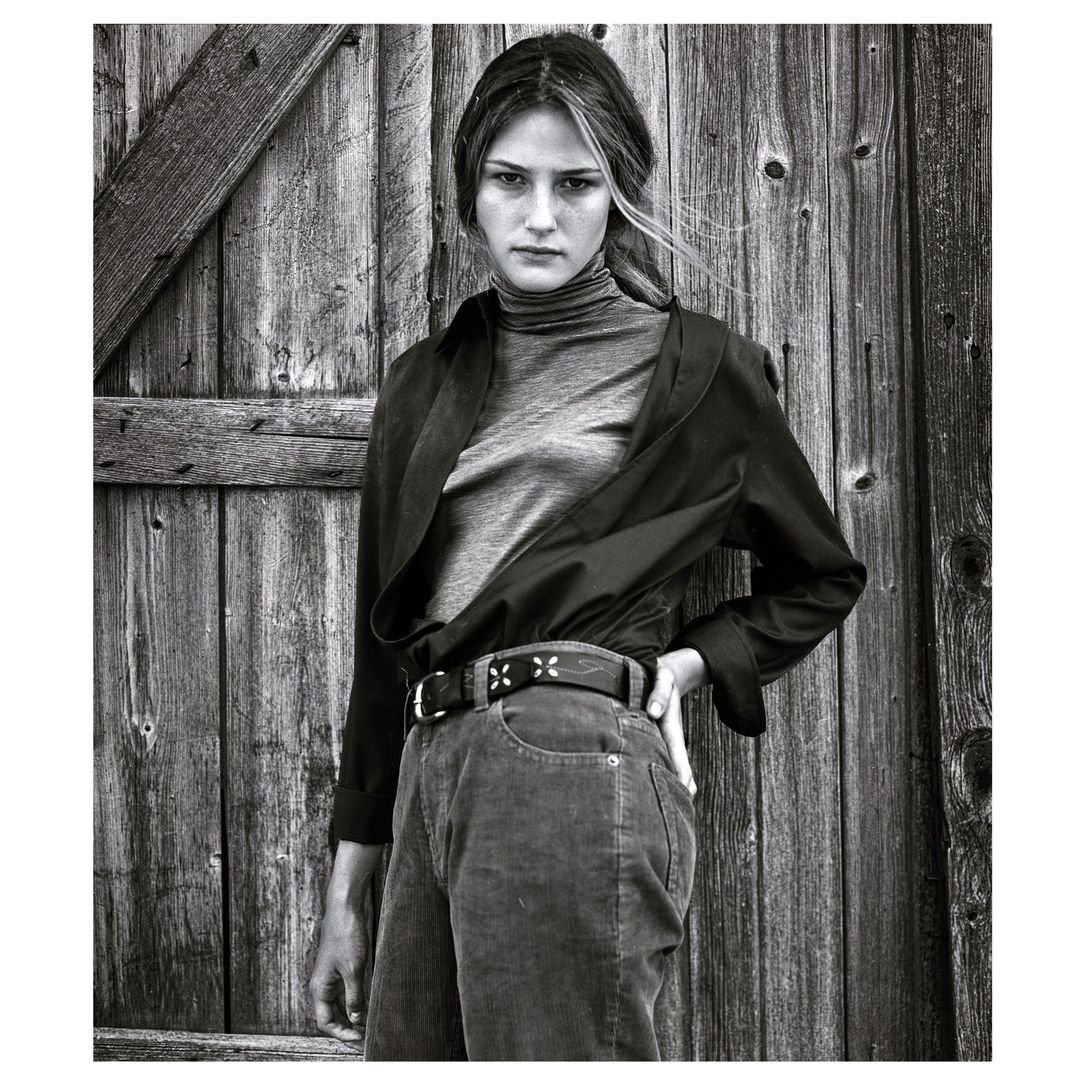 Black and white portrait of a woman standing in front of a wooden door wearing a turtleneck, pants and a black, unbuttoned shirt.