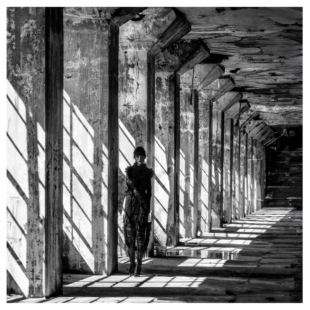 Black and white fashion shot of a woman in a textured dress standing amidst concrete pillars casting strong shadows in an urban environment.
