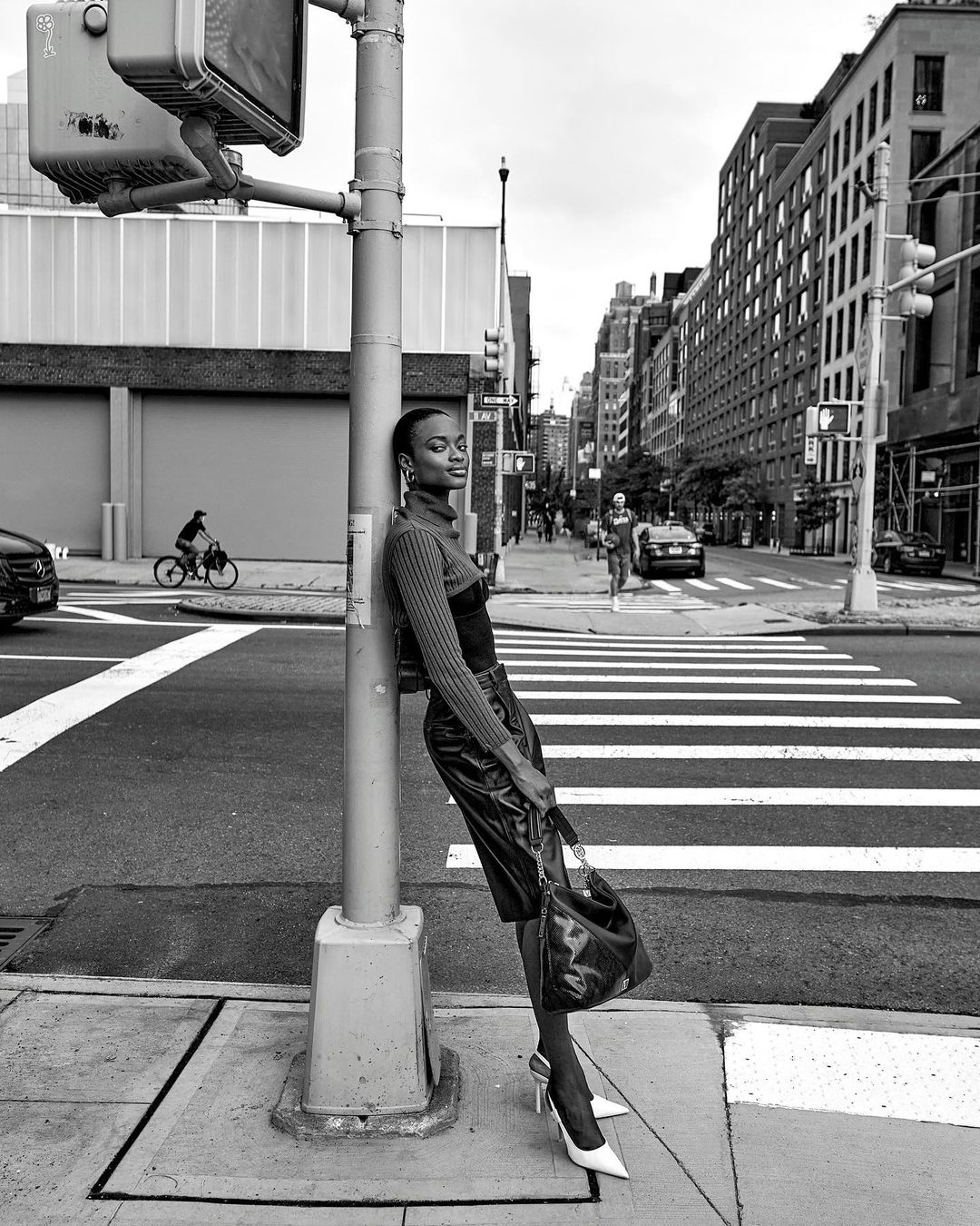 Black and white fashion shot of an adult woman leaning on a traffic pole in a city street, wearing heels and a leather skirt.