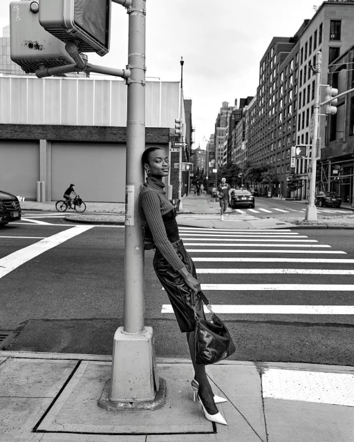 Black and white fashion shot of an adult woman leaning on a traffic pole in a city street, wearing heels and a leather skirt.