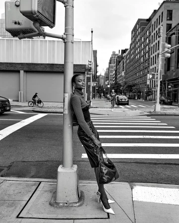 Black and white fashion shot of an adult woman leaning on a traffic pole in a city street, wearing heels and a leather skirt.