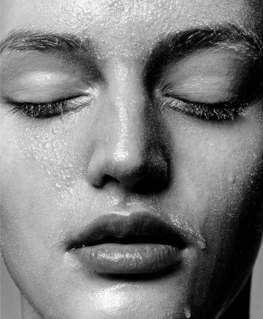 Close-up portrait of an adult female face covered in water droplets, showcasing dewy skin and a luminous, fresh complexion with closed eyes.